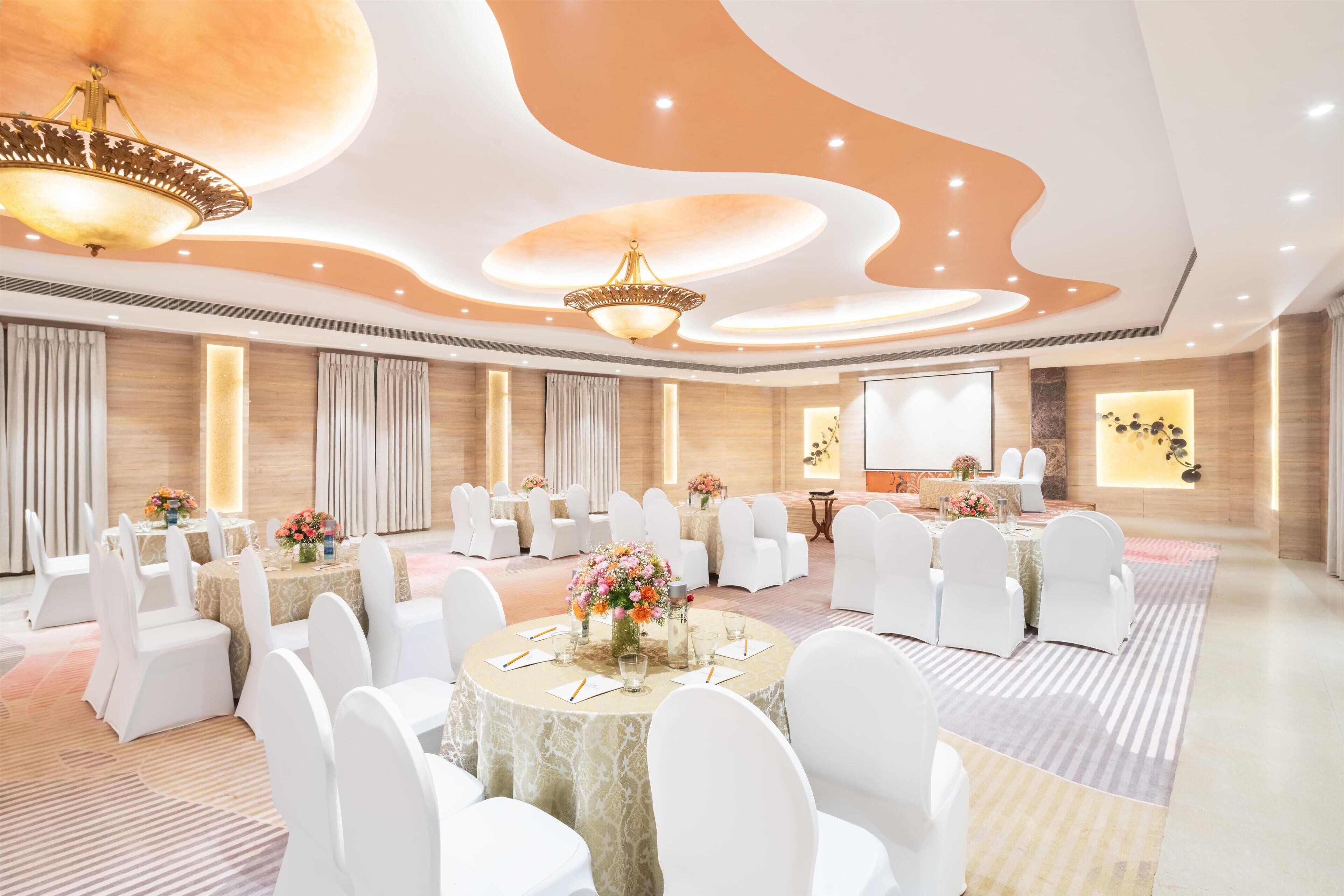 A spacious conference room with round tables, white chairs, and elegant chandeliers.