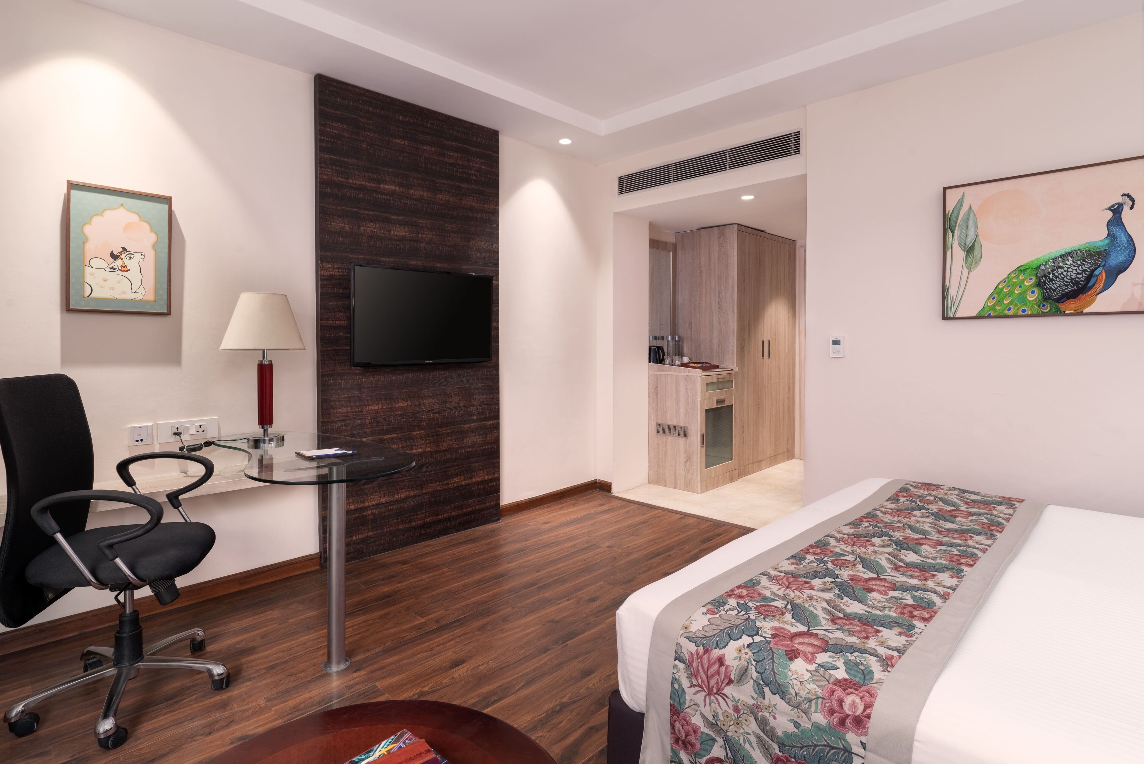Habitación de hotel con cama, escritorio con lámpara, silla, televisor y un cuadro de un pavo real.
