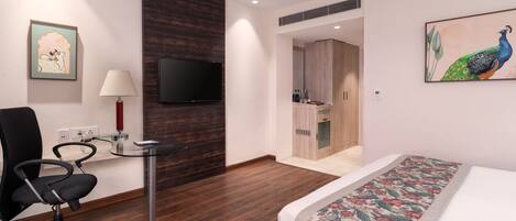 Habitación de hotel con cama, escritorio con lámpara, silla, televisor y un cuadro de un pavo real.
