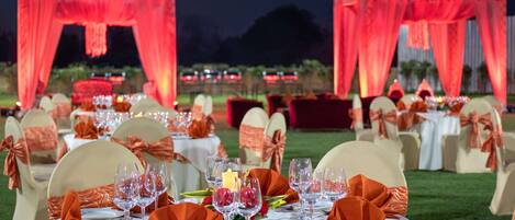 A formal outdoor dining setup with tables set for a meal, chairs with orange sashes, and red drapery.