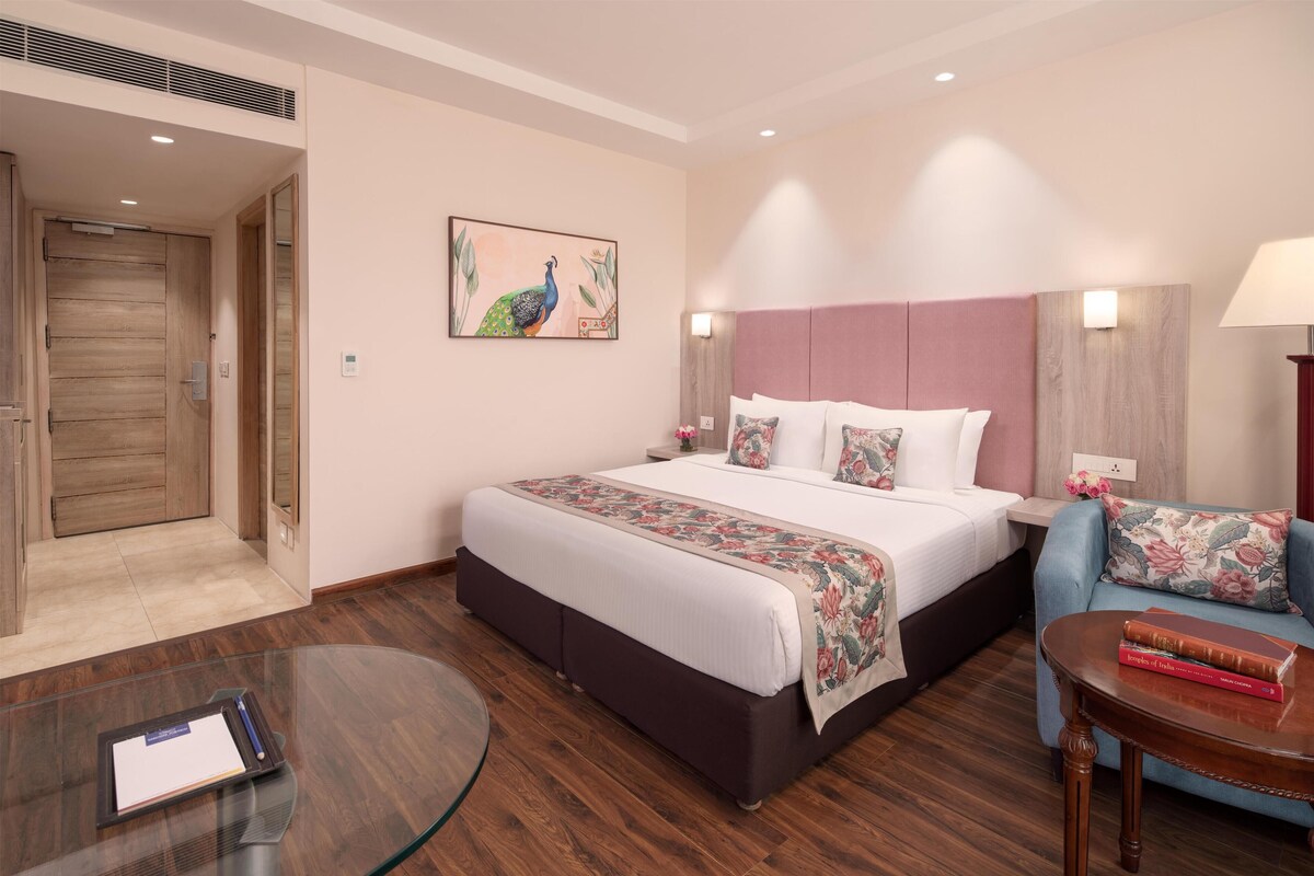 A hotel room with a large bed, a desk with a chair, a small table with books, and a door leading to another room.