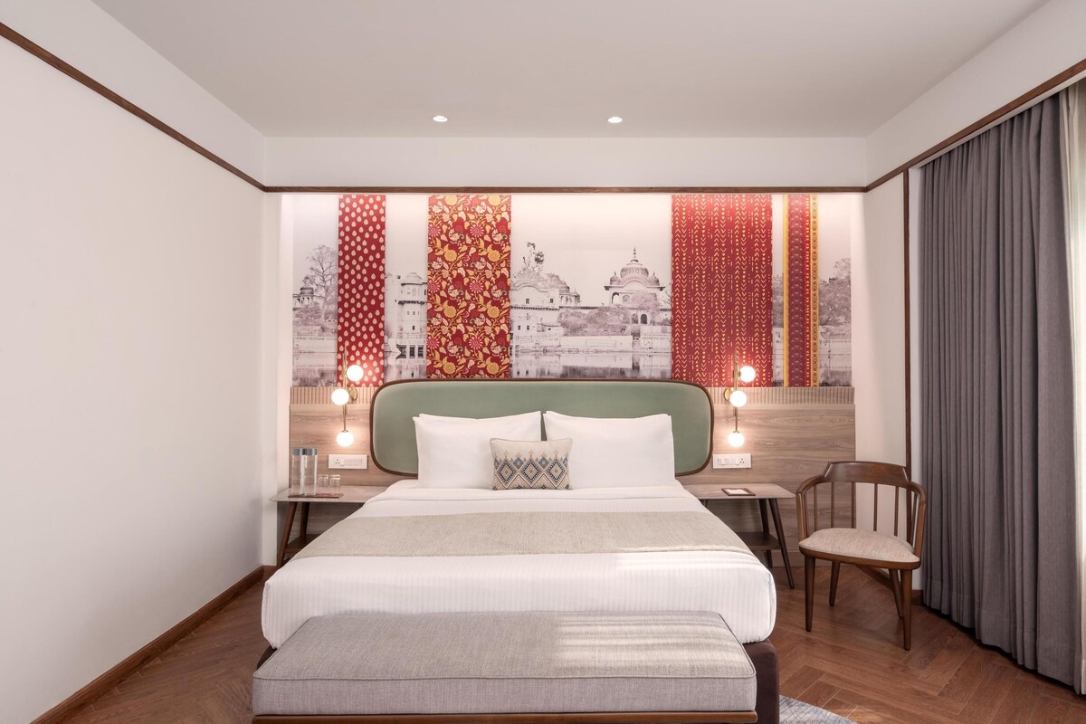 A modern hotel room with a large bed, a wooden headboard, a desk with a chair, and a view of a cityscape through patterned curtains.