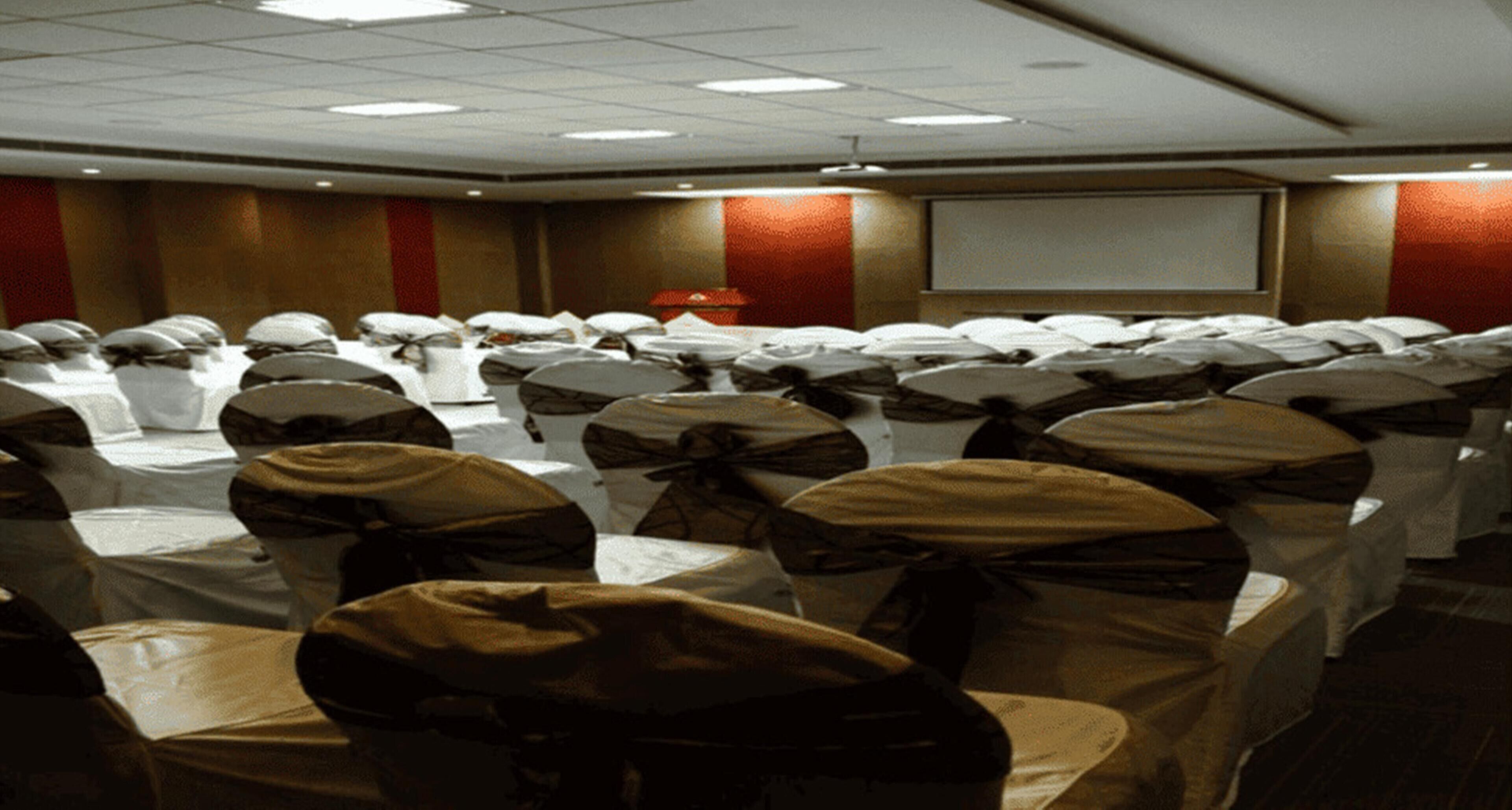 A conference room with rows of chairs covered in white tablecloths and brown sashes, a projector screen, and a red accent wall.
