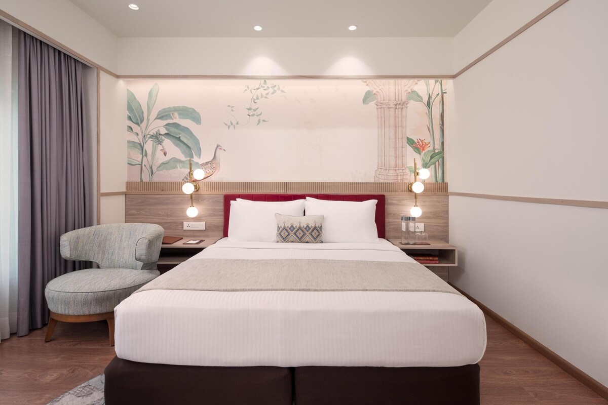 A hotel room with a large bed, a chair, a nightstand with a lamp, and a wall mural.