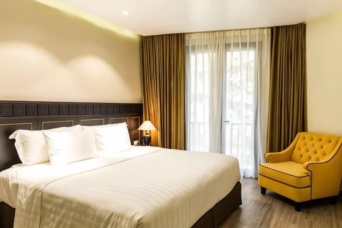 A hotel room with a large bed, a bedside lamp, a yellow armchair, and a window with curtains.