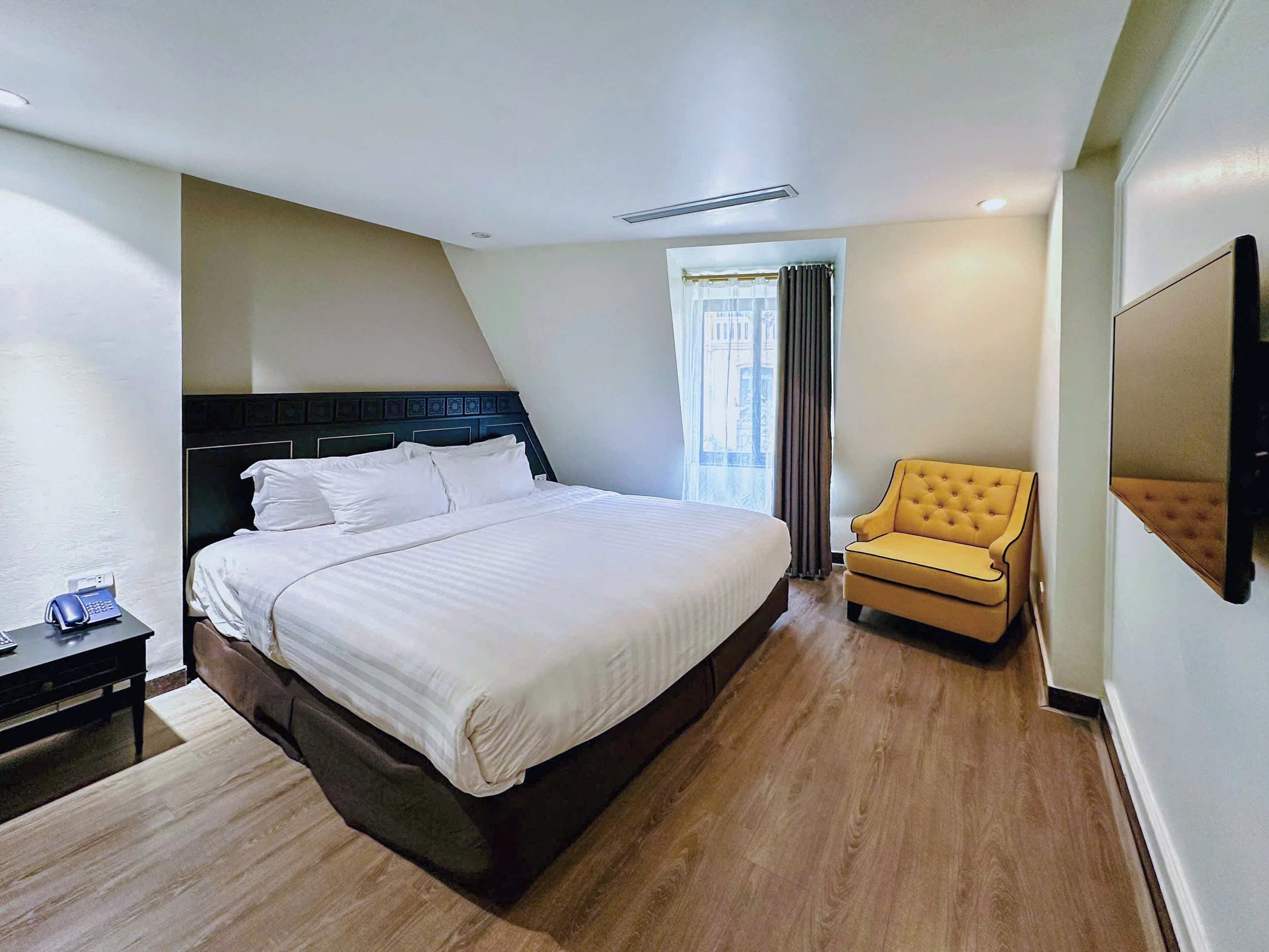 A hotel room with a large bed, a bedside table with a phone, a chair, and a TV mounted on the wall.