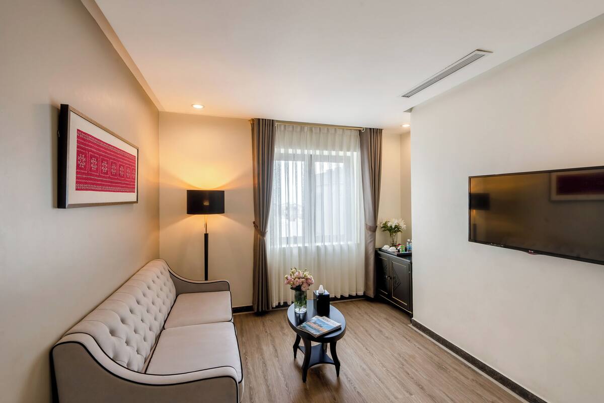 A modern living room with a sofa, a small table with a vase of flowers, a lamp, and a TV mounted on the wall.