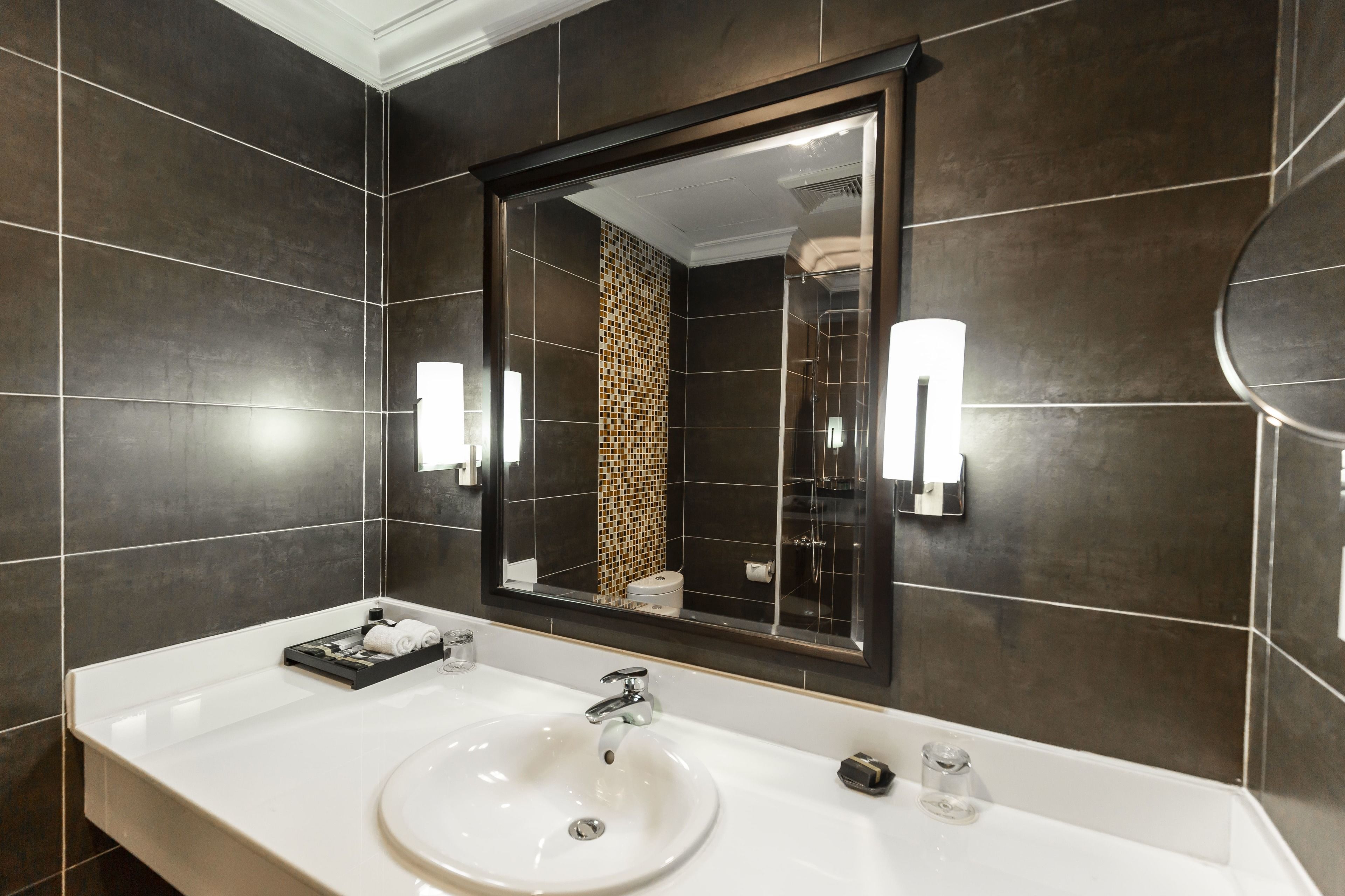A modern bathroom with a large mirror, dark tiled walls, a white sink, and two wall-mounted lights.