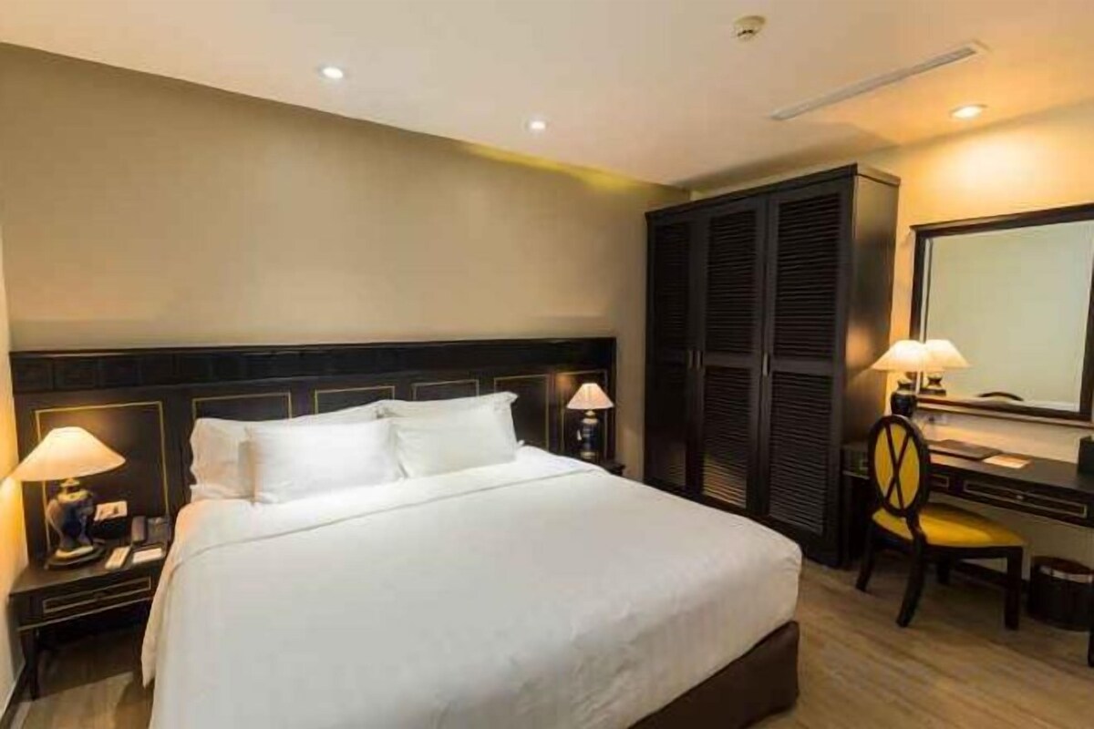 A hotel room with a large bed, bedside lamps, a desk with a chair, a mirror, and a wardrobe.