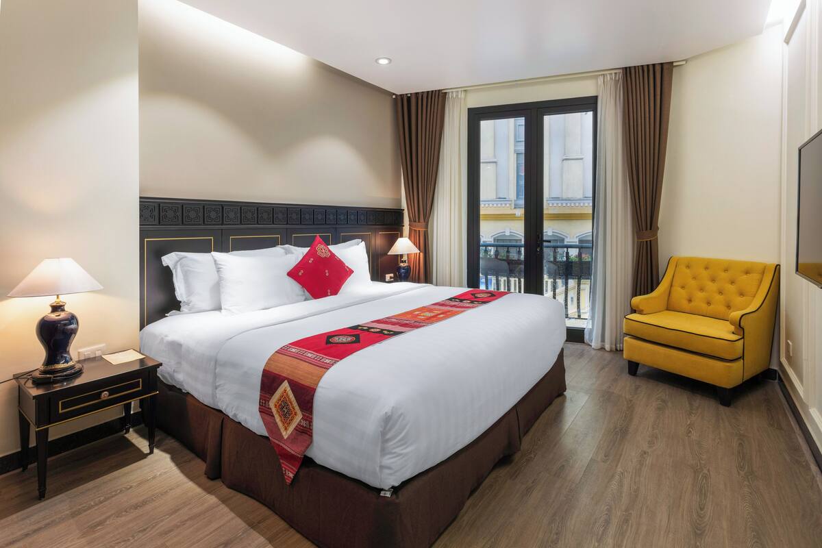 A hotel room with a large bed, a yellow armchair, a nightstand with a lamp, and a view of the outside through a sliding door.