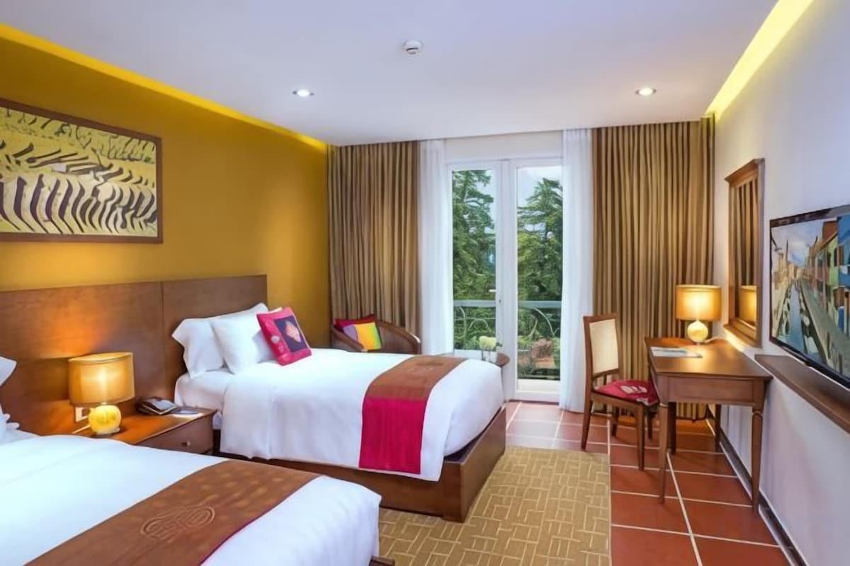 A hotel room with two beds, a desk, a chair, a lamp, and a view of the outdoors.