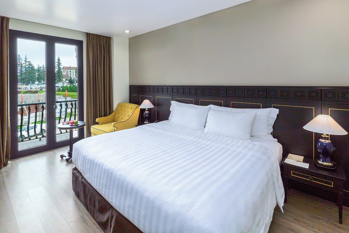 A hotel room with a large bed, a small table, a chair, and a balcony with a view of the street.