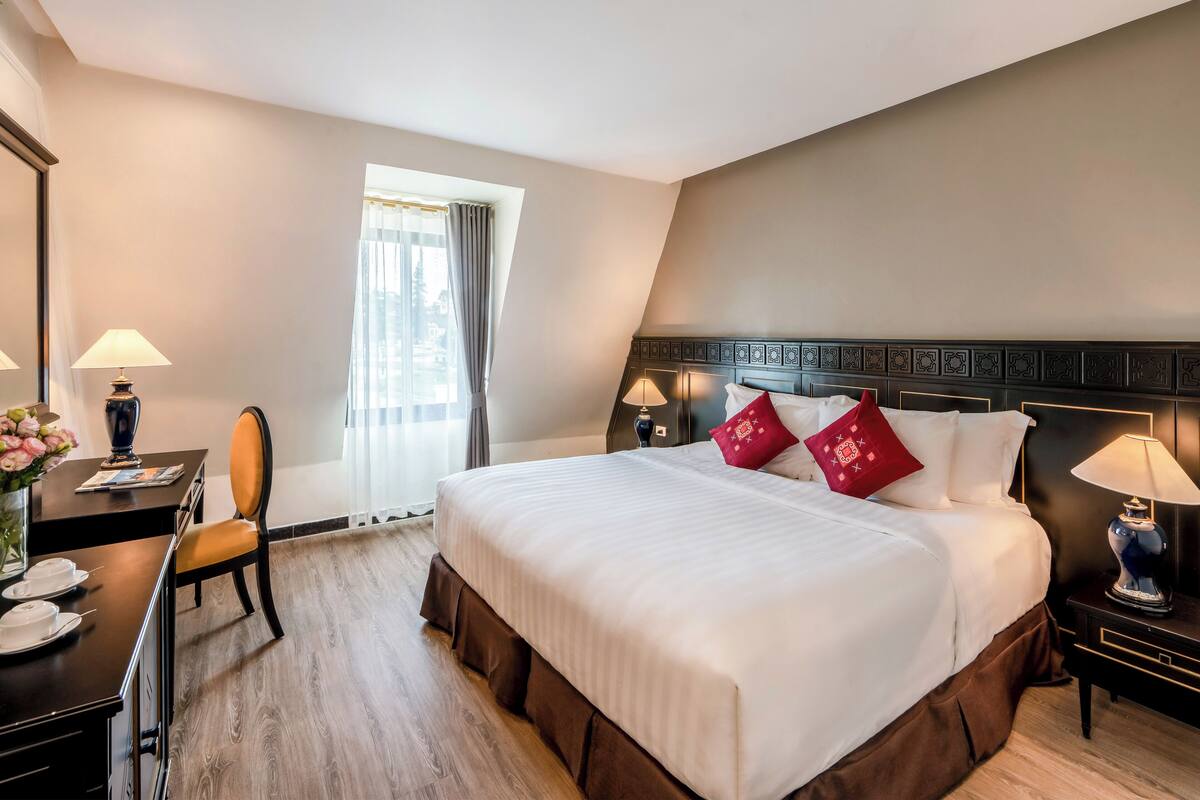 A hotel room with a large bed, a desk with a chair, a nightstand with a lamp, and a window with curtains.