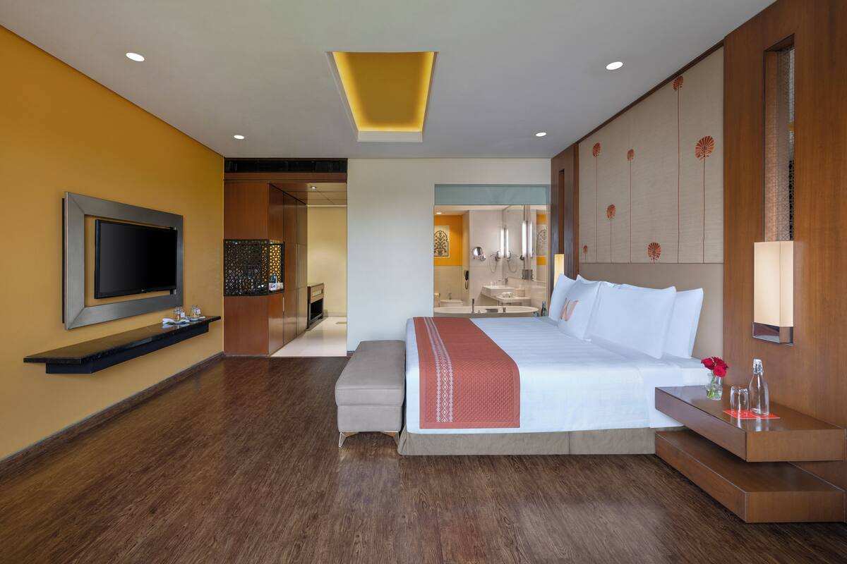 Een moderne hotelkamer met een groot bed, een aan de muur bevestigde televisie, een nachtkastje met een lamp en een klein bankje.