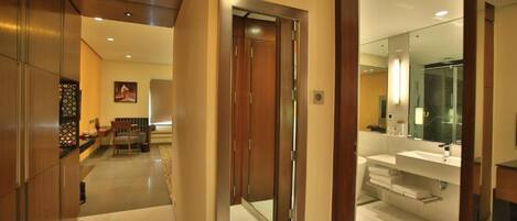 Un pasillo moderno de hotel con puerta de madera, un área de comedor con mesa y sillas, y un baño con un espejo grande y lavabo.