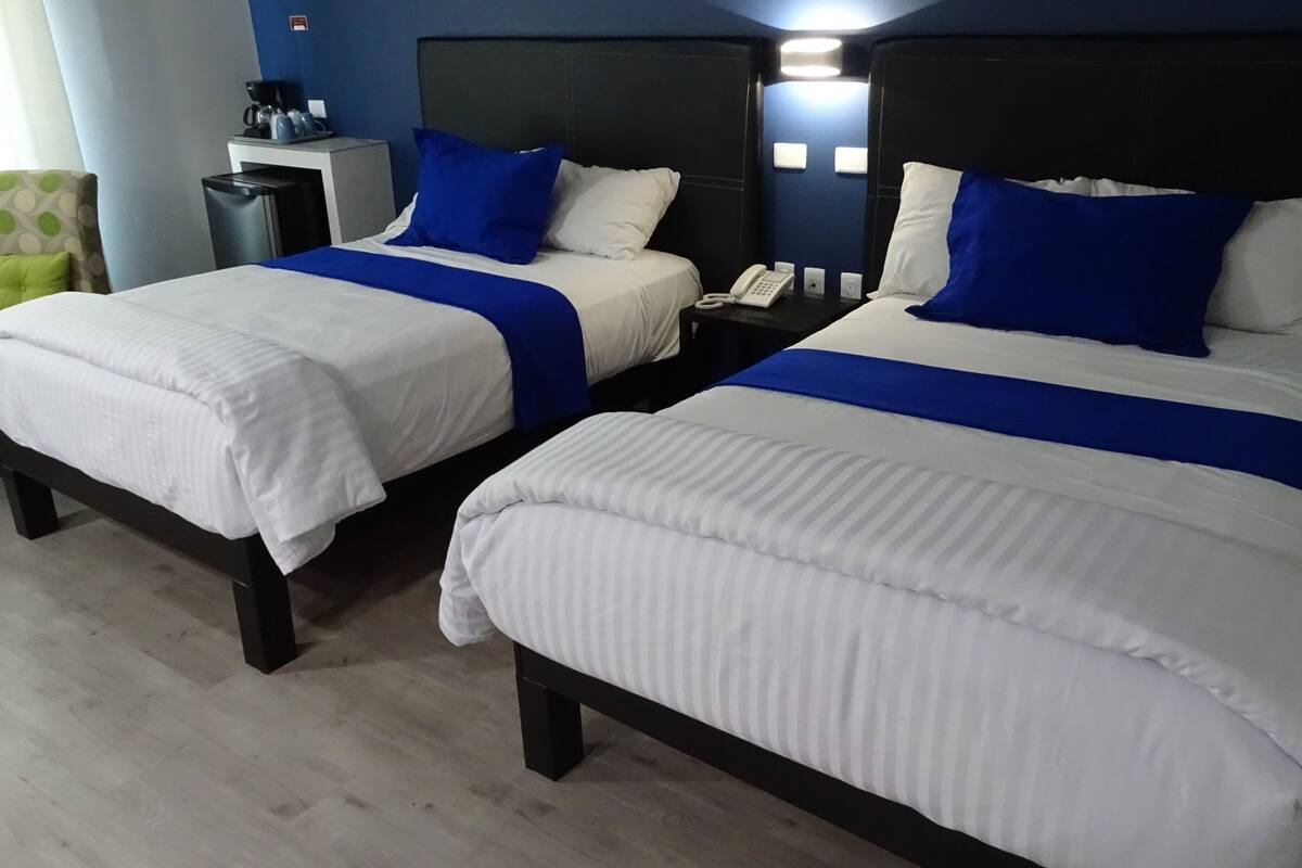 A hotel room with two beds, each with white bedding and blue accents, a bedside table with a phone, a chair with a green polka dot cushion, and a coffee machine on a small table.