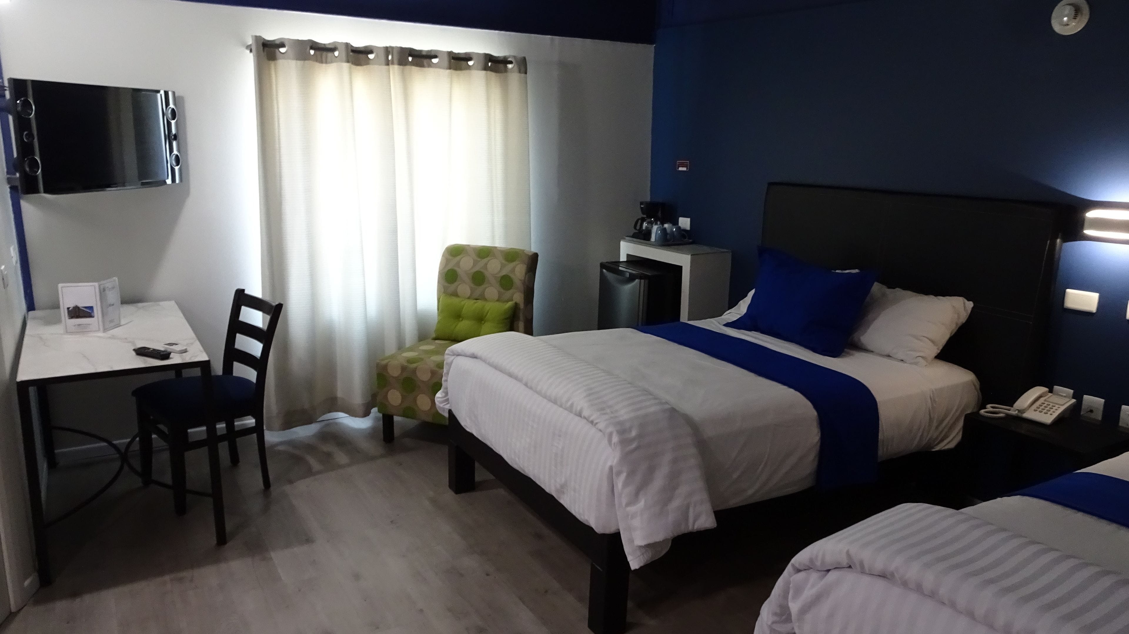 Una habitación de hotel con cama, escritorio, silla, televisor y ventana con cortinas.