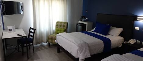 Una habitación de hotel con cama, escritorio, silla, televisor y ventana con cortinas.