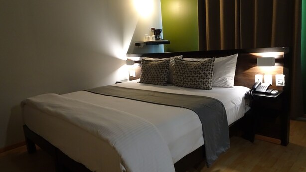 Cama bem arrumada com edredom branco e cinza, duas almofadas estampadas e mesa de cabeceira com telefone e abajur.