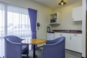 Hotelkamer met een eethoek met twee stoelen en een klein tafeltje, een kitchenette met een magnetron, en een uitzicht door een raam met dunne gordijnen.