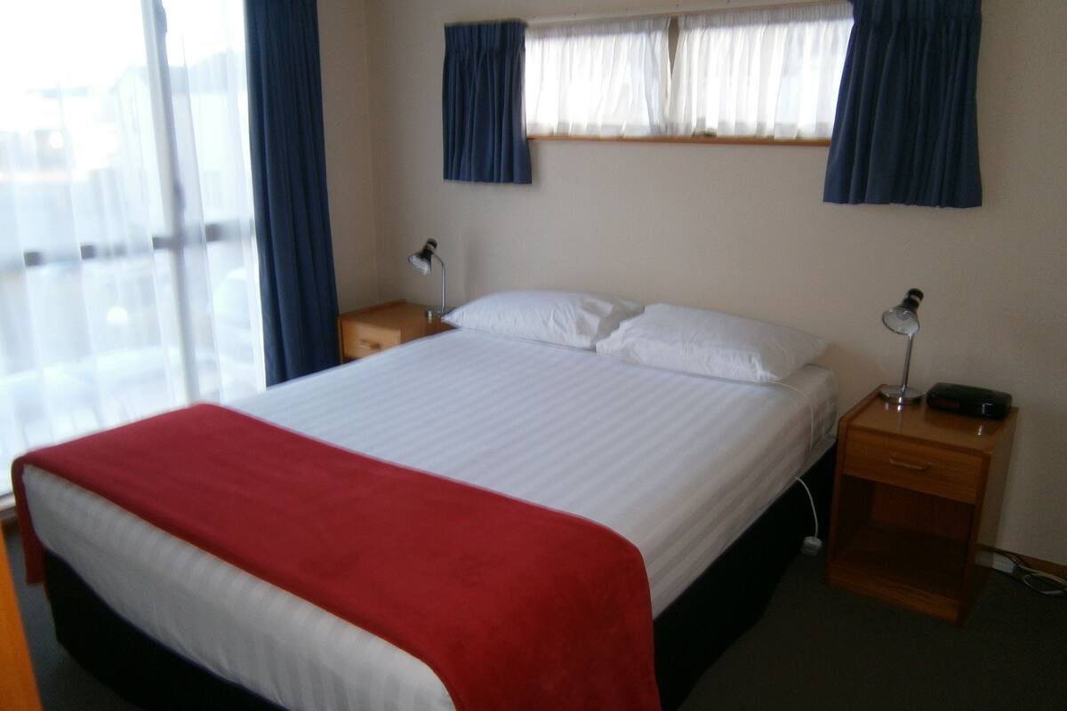 A neatly made bed with a red blanket, two bedside tables with lamps, and a window with blue curtains.