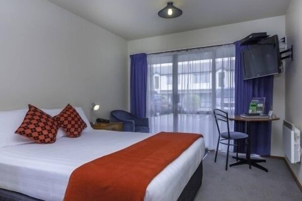 A hotel room with a bed, two pillows, a desk with a chair, a television, and a window with curtains.