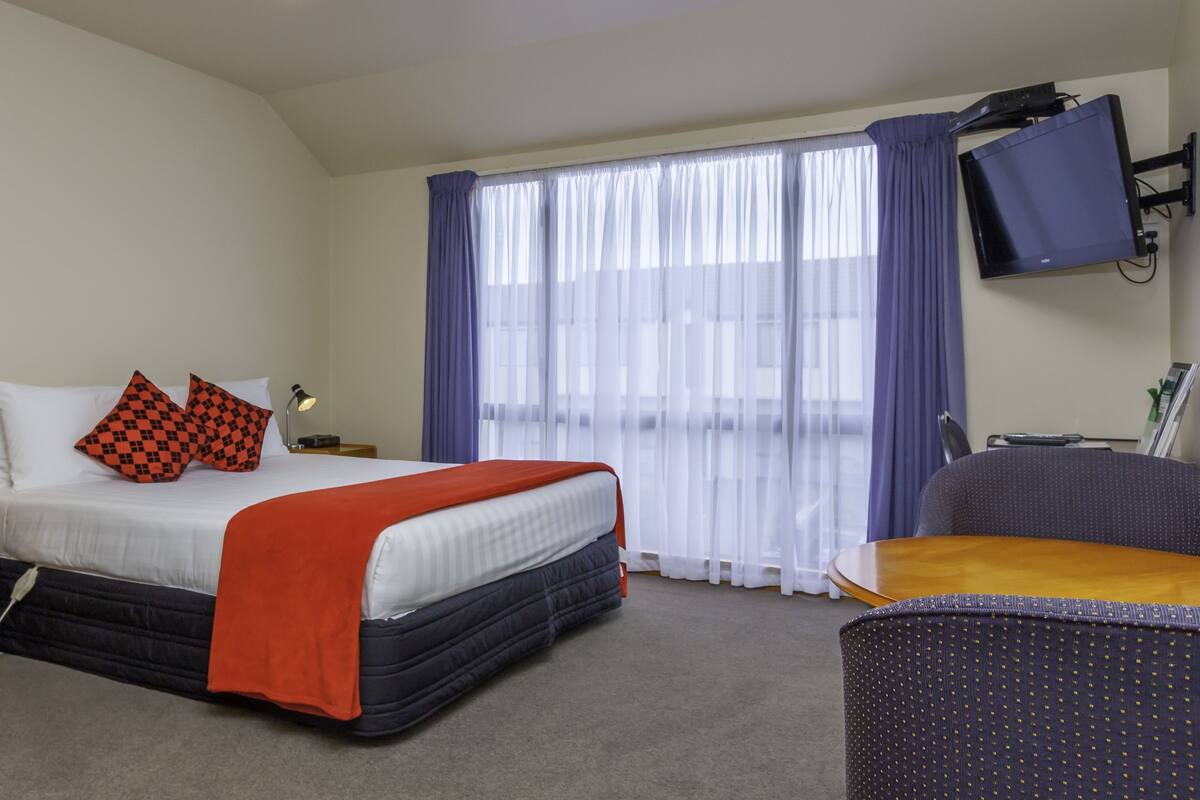 A hotel room with a bed, bedside tables, a desk, a chair, a TV, and a window with curtains.