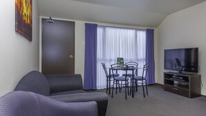 A hotel room with a sofa, a dining table with chairs, a television, and a window with curtains. - Deans Bush Motel (Christchurch)