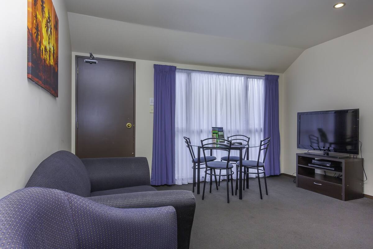 A hotel room with a sofa, a dining table with chairs, a television, and a window with curtains.