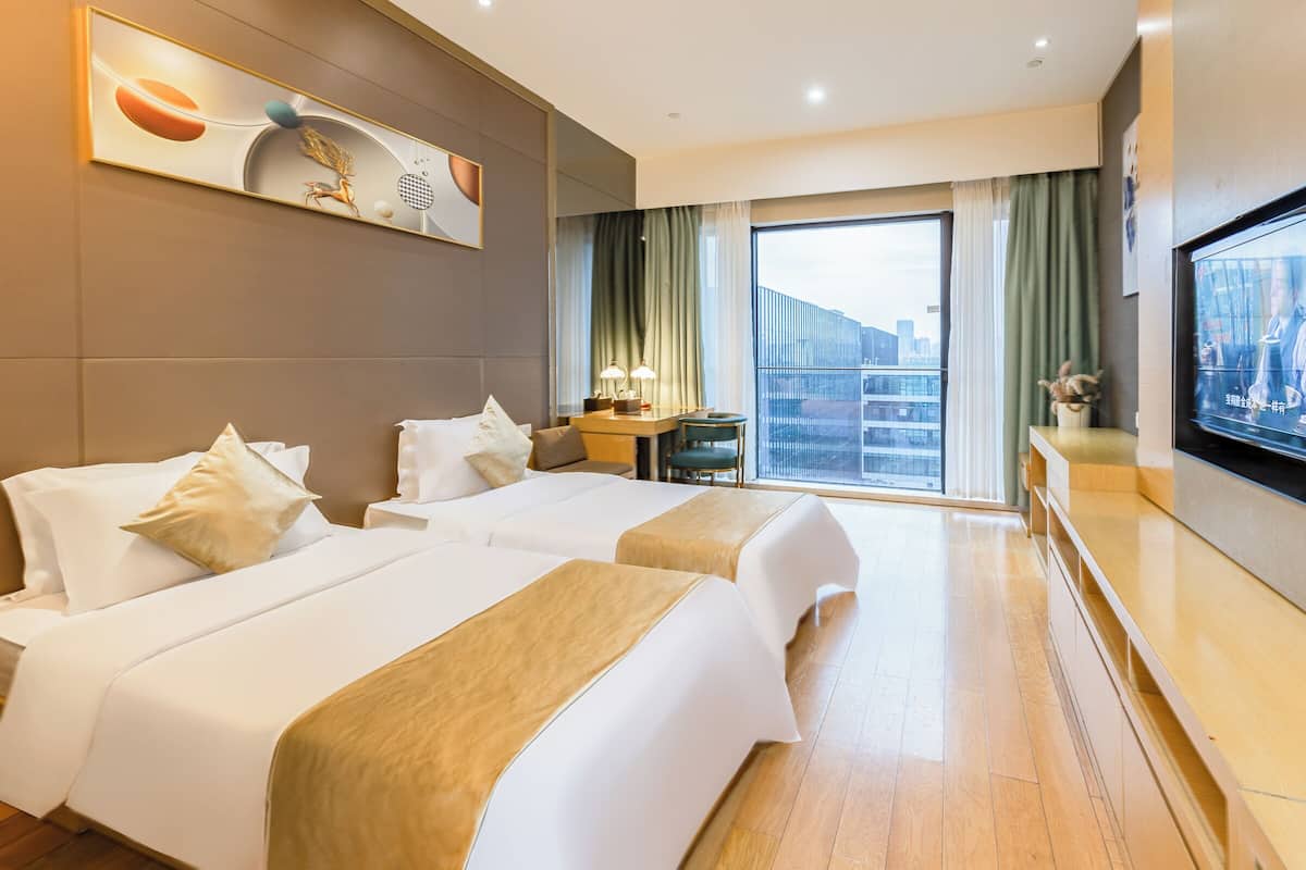 Habitación de hotel con dos camas, un escritorio con silla, televisión y un ventanal con vistas a edificios.