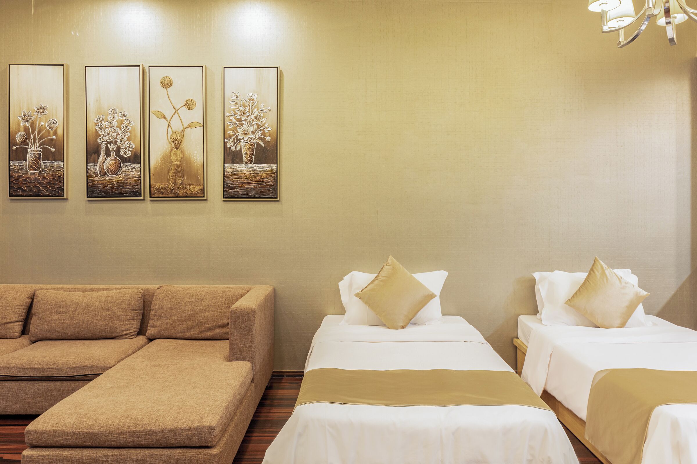 A hotel room with a sofa, two beds with gold accents, and three framed floral paintings on the wall.