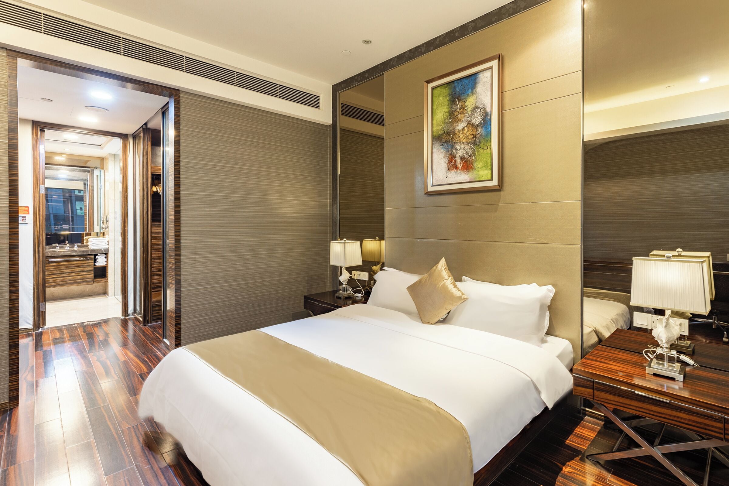 A hotel room with a large bed, wooden floor, a painting on the wall, and a nightstand with a lamp.