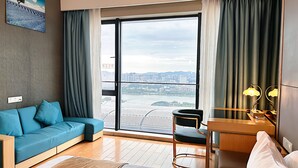 A modern hotel room with a large sliding glass door to a city view, a blue sofa, a wooden desk with lamps, and a chair. - Xingyi International Hotel Poly Store (Guangzhou)