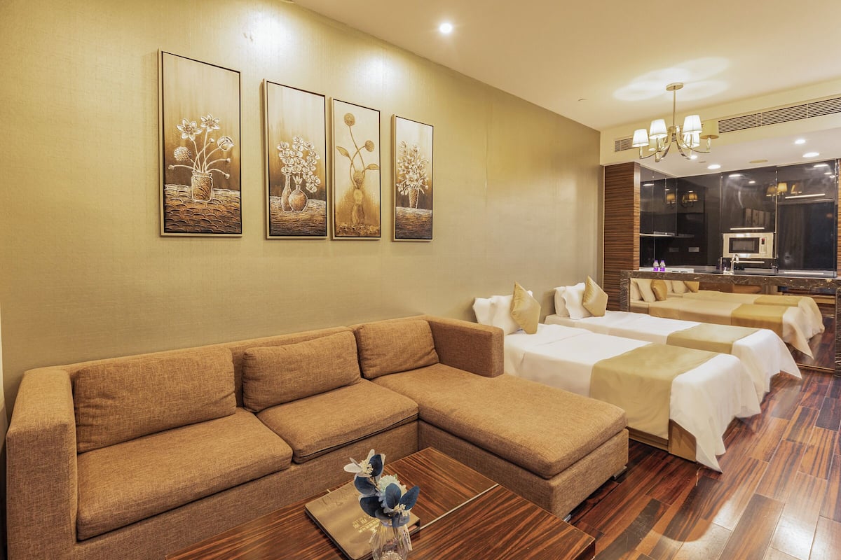 Habitación de hotel con un sofá seccional marrón, dos camas individuales, una mesa de centro de madera con un jarrón, y tres cuadros colgados en la pared.