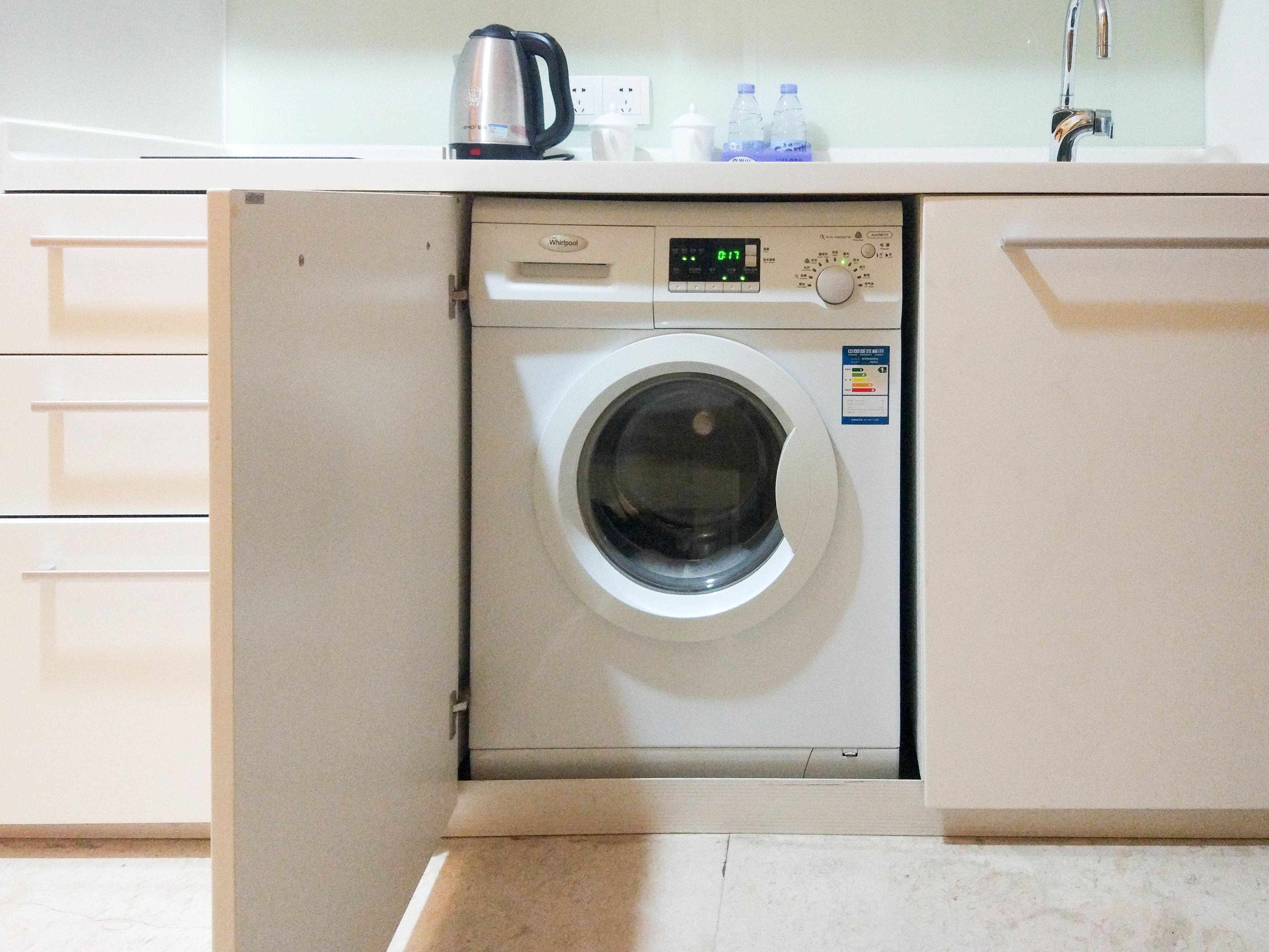 Máquina de lavar Whirlpool branca e compacta numa cozinha com bancada, chaleira e duas garrafas de água.