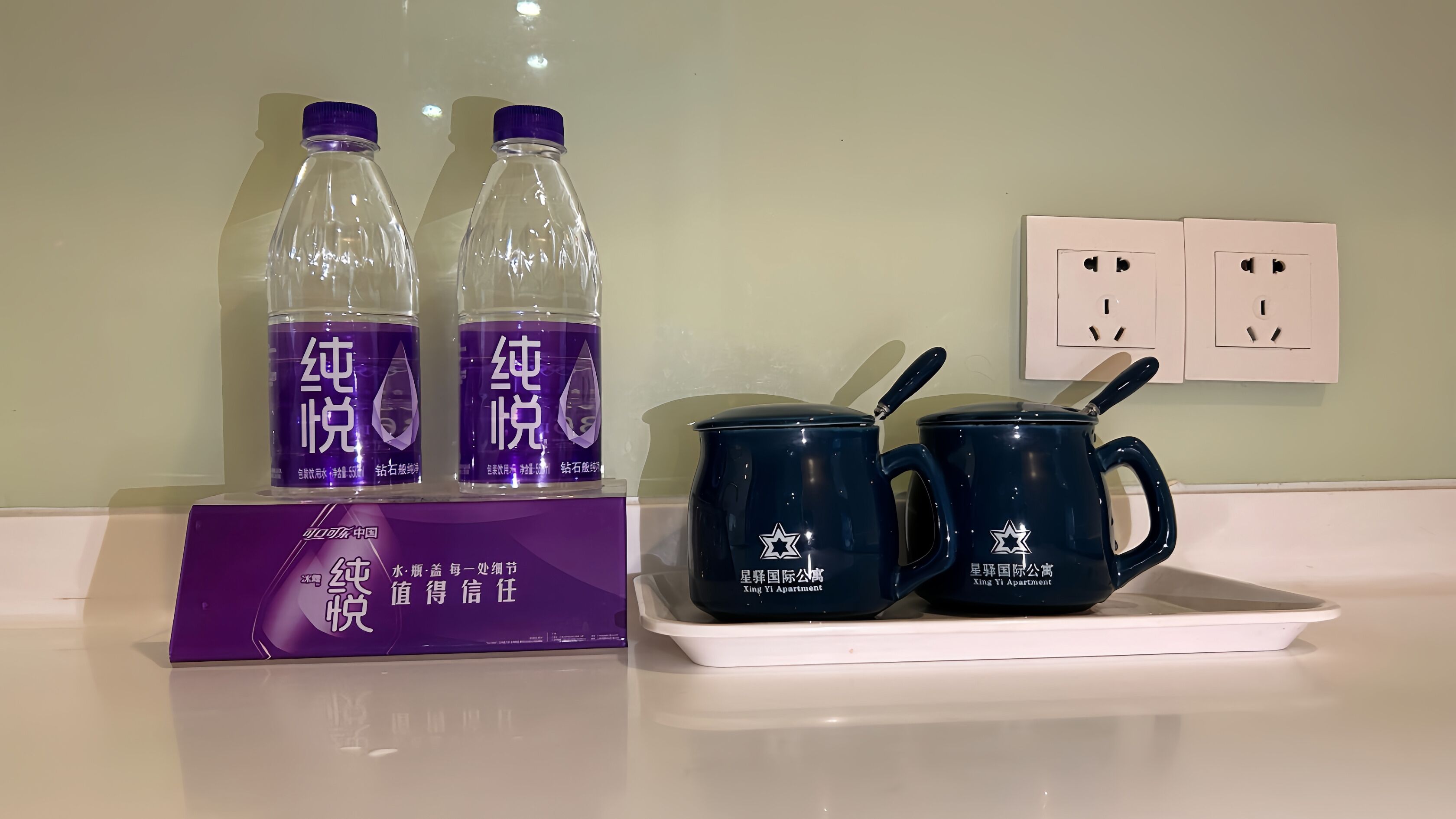 Habitación de hotel con un letrero morado, dos botellas de agua y dos tazas azul oscuro sobre una bandeja.