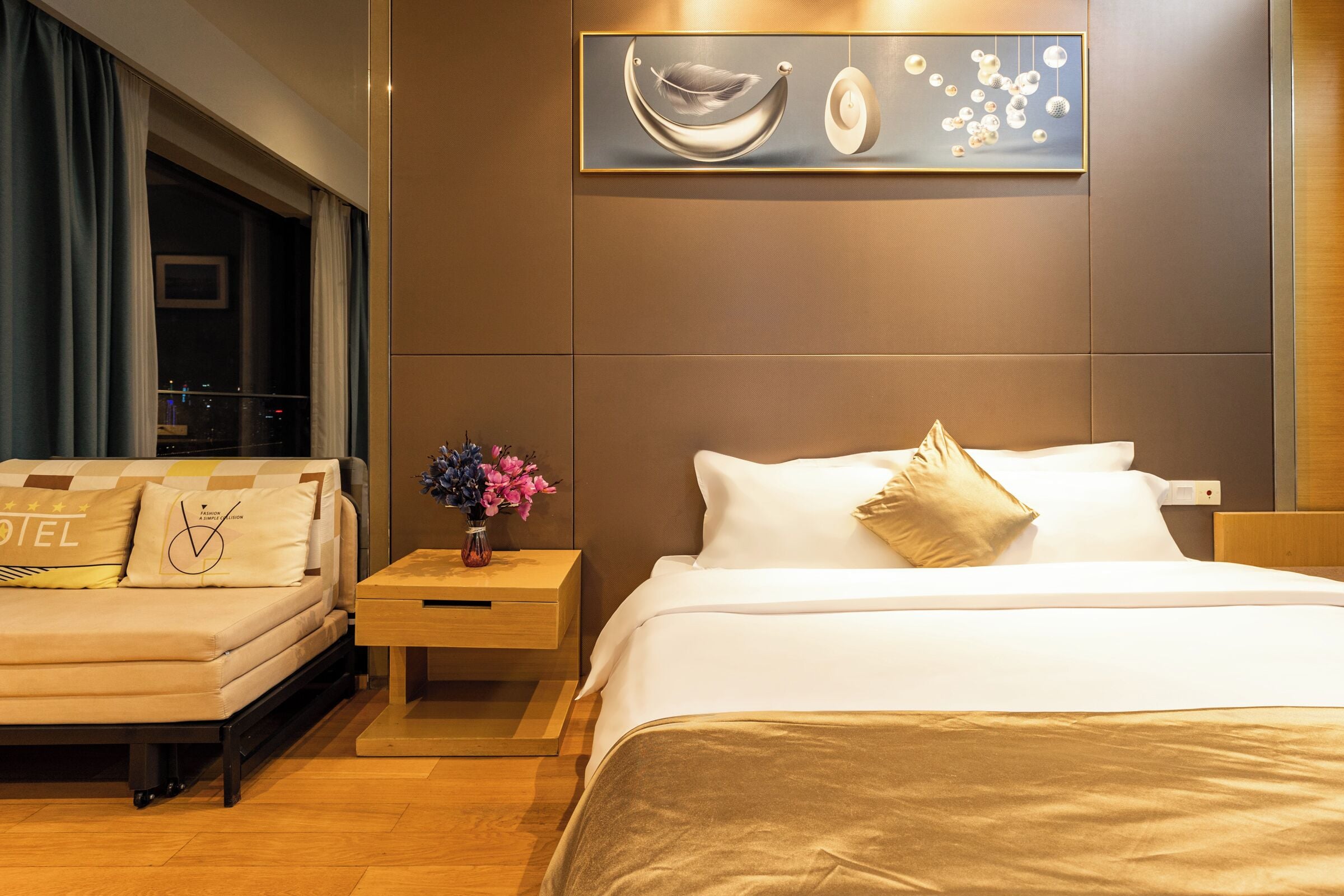 A hotel room with a bed, a sofa, a nightstand with a vase of flowers, and a wall-mounted artwork.