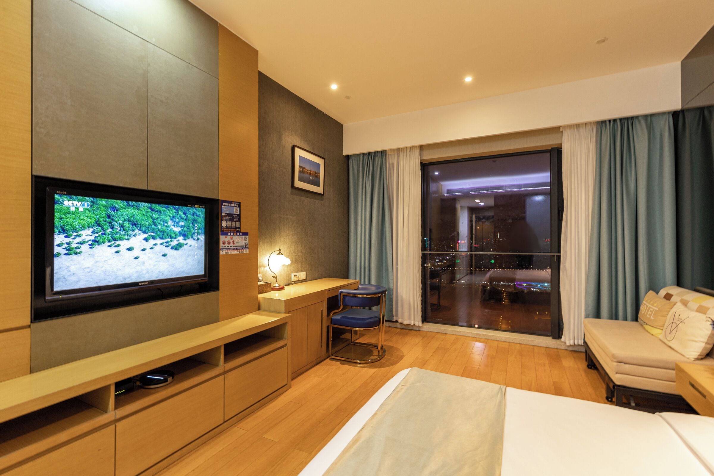 A hotel room with a TV, a desk, a chair, a bed, a sofa, and a large window with curtains.