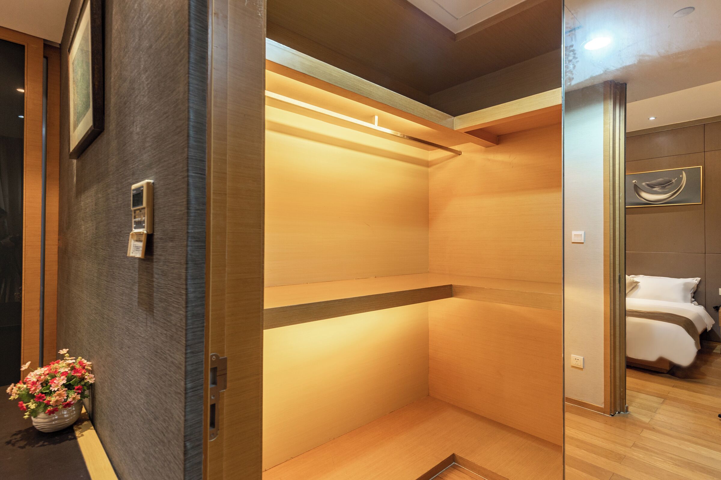 A hotel room with a wooden floor, a bed with white bedding, a small bedside table, and a wall-mounted shelf.