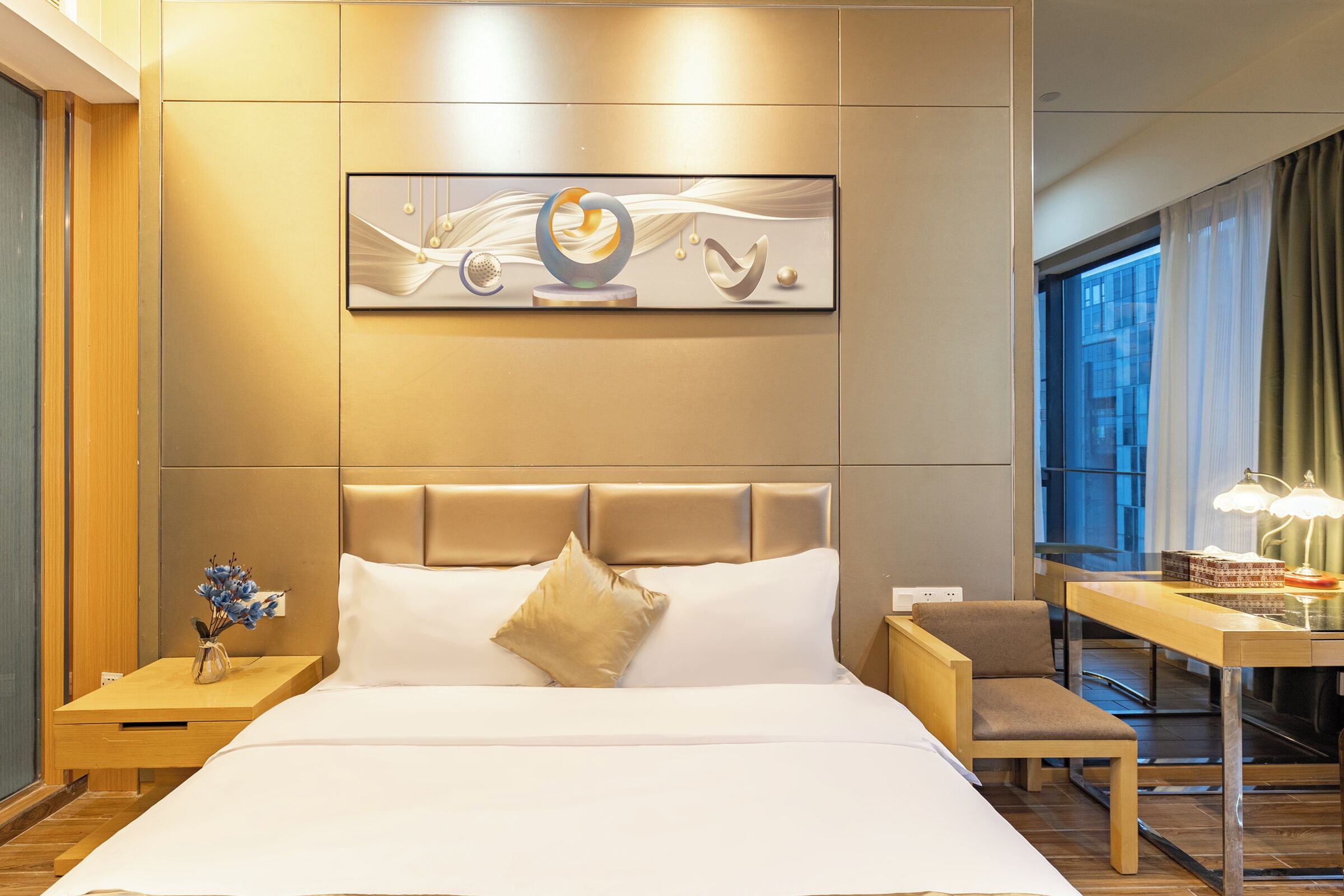 A modern hotel room with a large bed, a bedside table with a vase of flowers, a desk with a lamp, and a large abstract painting on the wall.