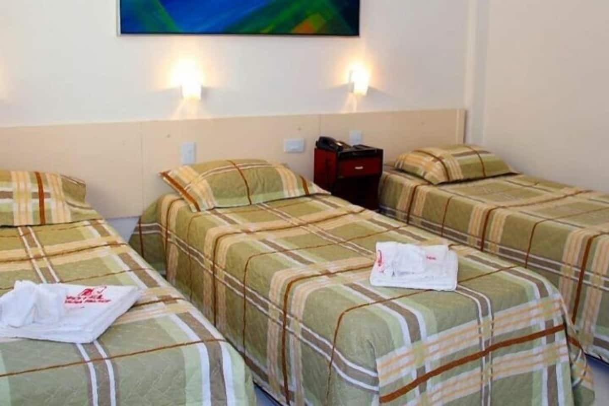 Ein Hotelzimmer mit drei Betten, jedes mit einer karierten Tagesdecke, einem roten Koffer auf einem Bett und einem weißen Handtuch auf einem anderen. Über den Betten hängt ein Gemälde an der Wand.