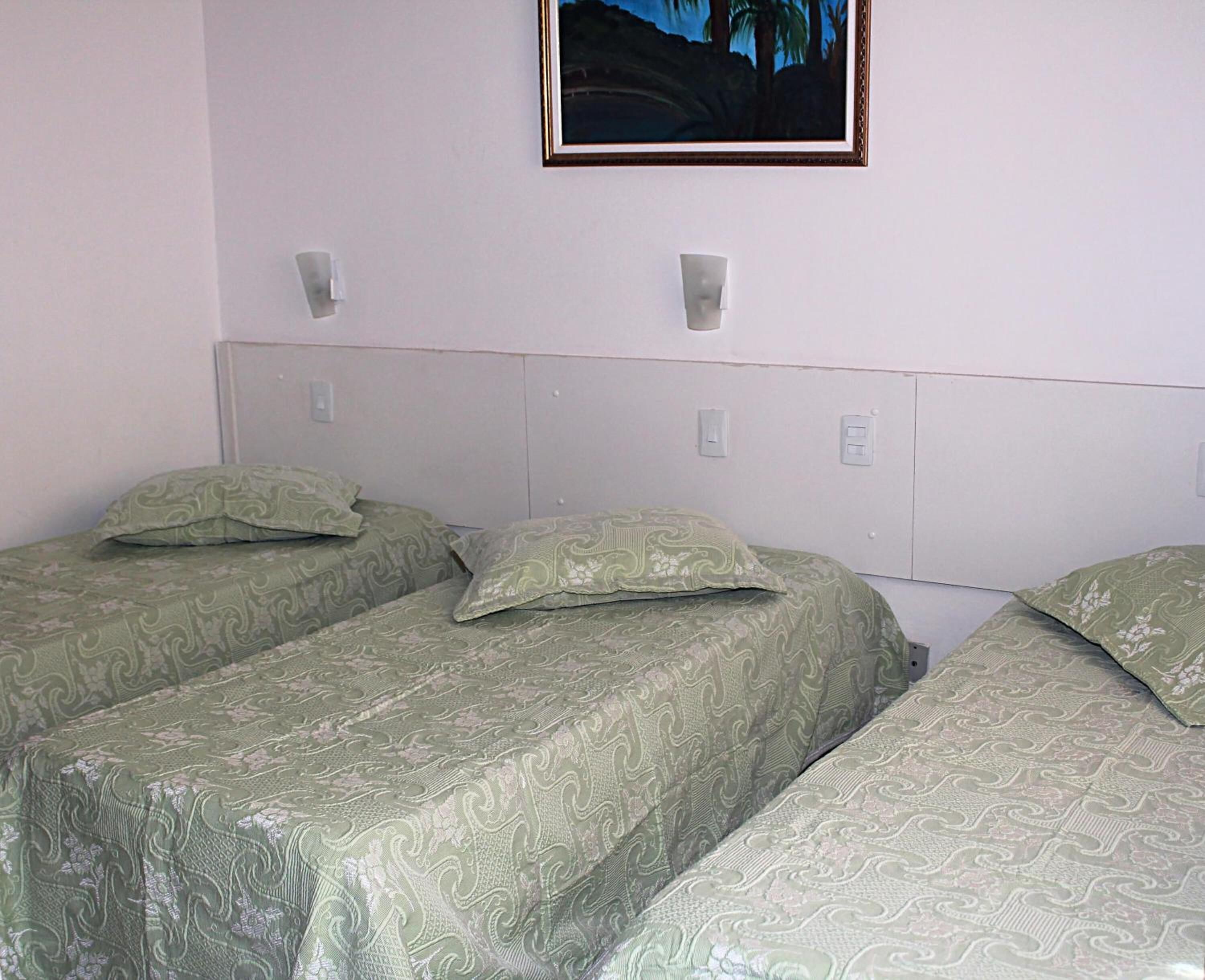 Une chambre avec trois lits simples, chacun recouvert d’une couverture verte à motifs et agrémenté d’oreillers assortis. Un tableau encadré est accroché au mur, au-dessus des lits.