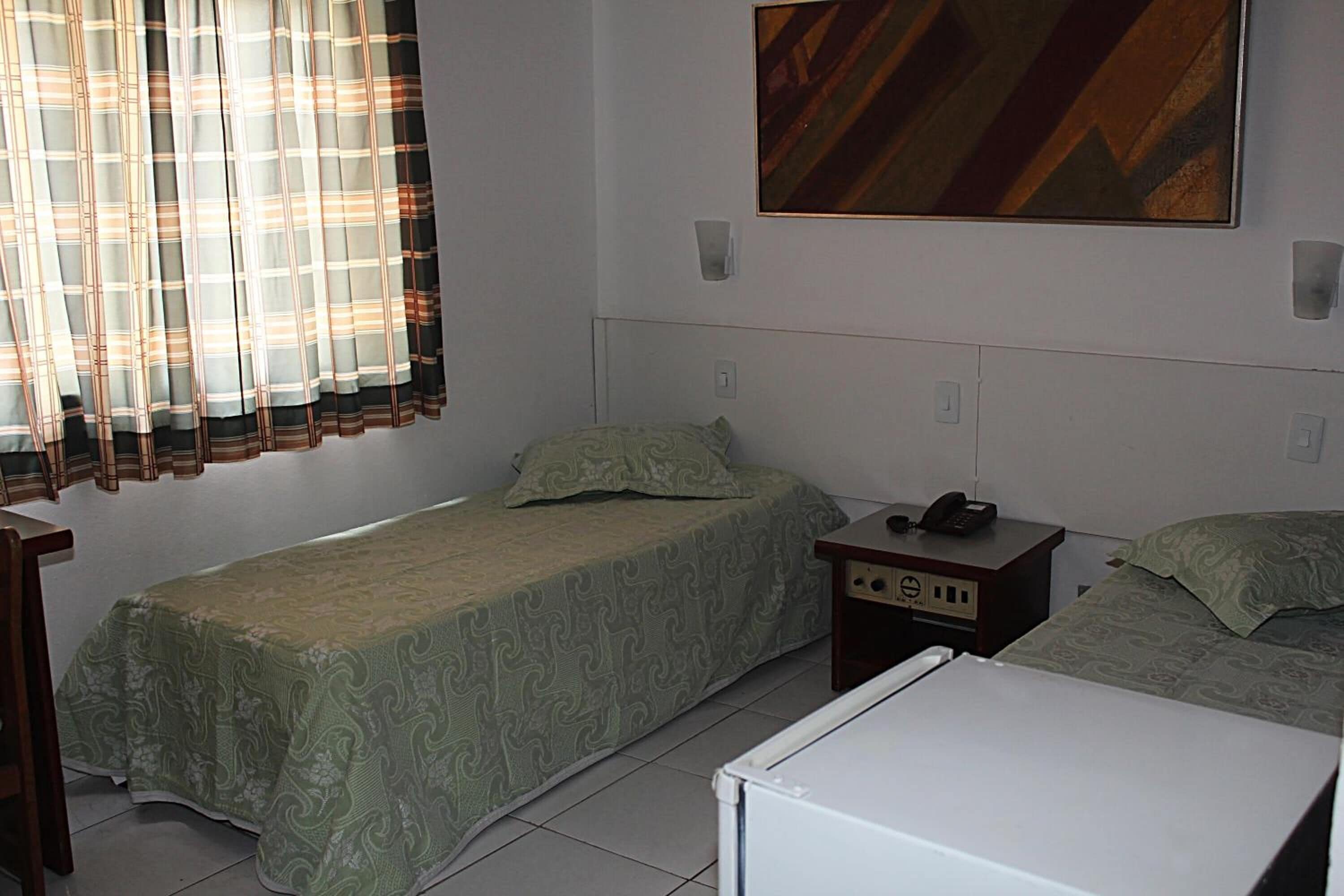 A small, simple room with two single beds, a desk, a chair, a telephone, and a refrigerator.