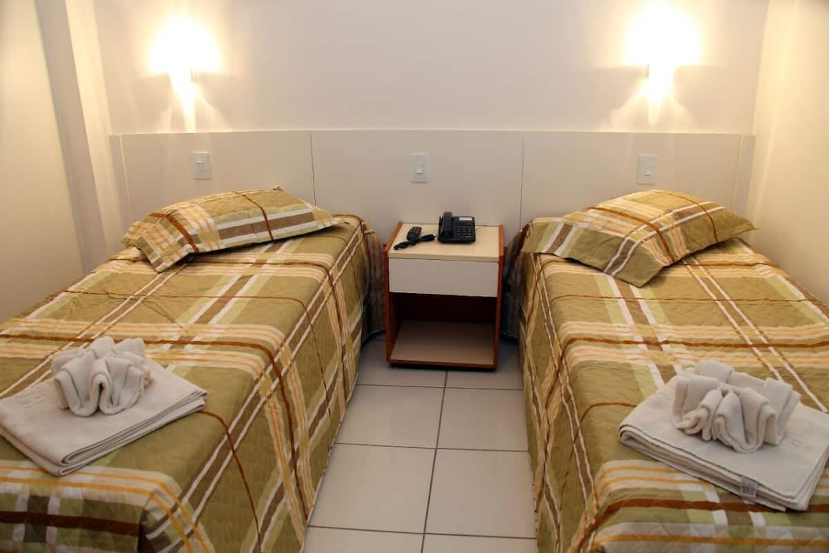Ein Hotelzimmer mit zwei Einzelbetten, jeweils mit Kissen und Handtuch. Zwischen den Betten steht ein Nachttisch mit einem Telefon.