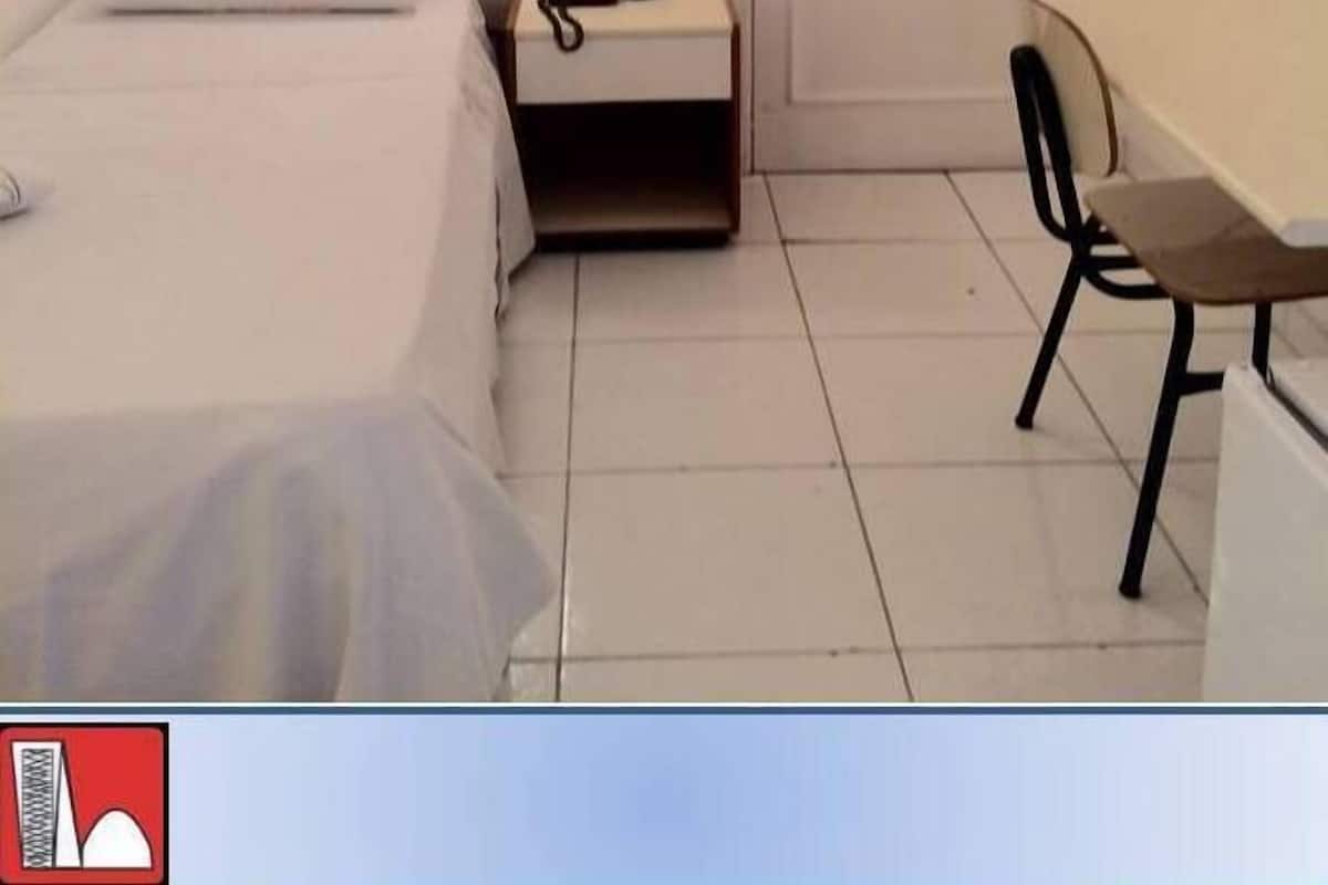 Ein kleines, schlichtes Zimmer mit einem Bett, einem Nachttisch mit Lampe und einem Schreibtisch mit Stuhl.