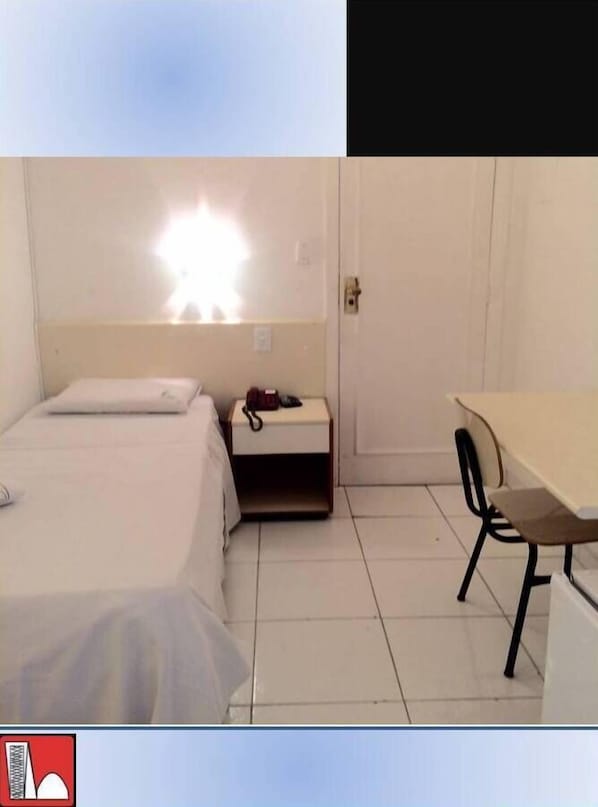 A small, simple room with a bed, a bedside table with a lamp, and a desk with a chair. - Hotel Pampulha (Belo Horizonte)