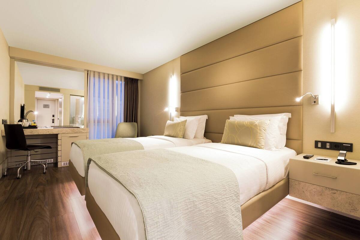 Une chambre d’hôtel avec deux lits, un bureau et une chaise. On y trouve un miroir, une table de chevet et une lampe.