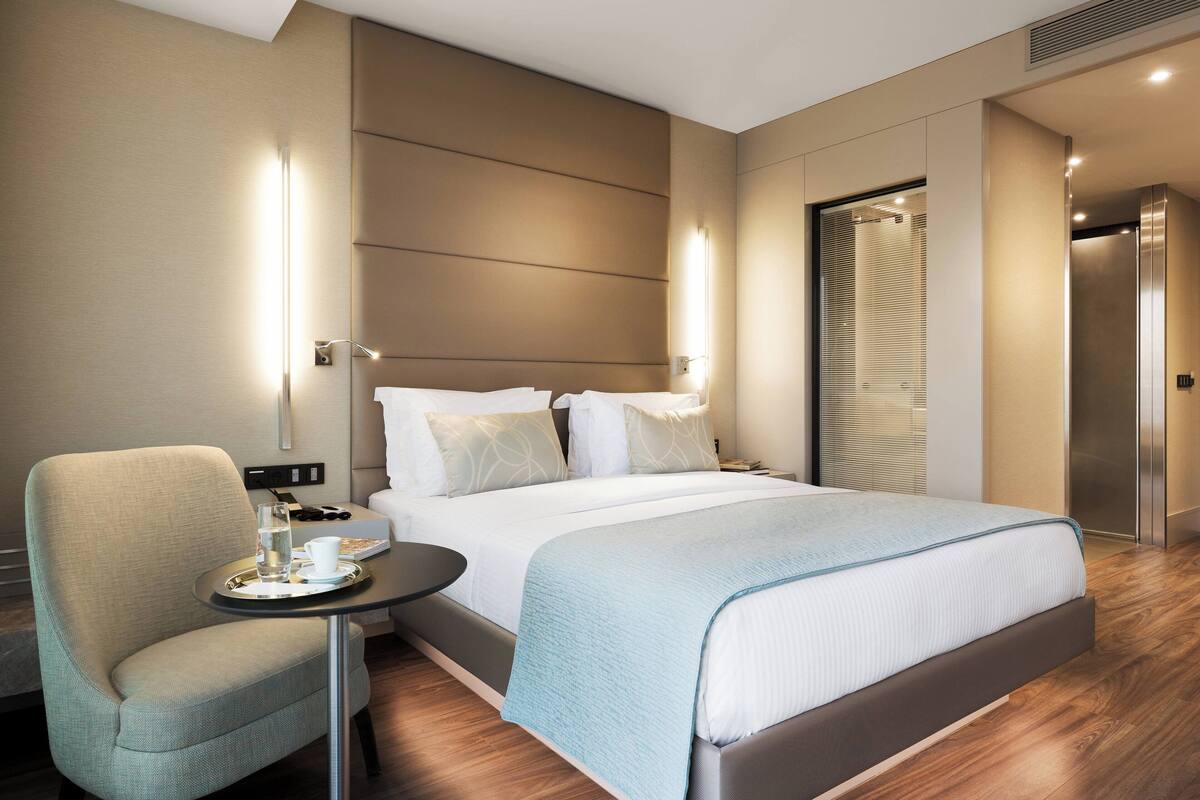 Une chambre d’hôtel moderne avec un grand lit, une petite table avec un plateau, une chaise et une salle de bain équipée d’un miroir et d’un porte-serviettes.