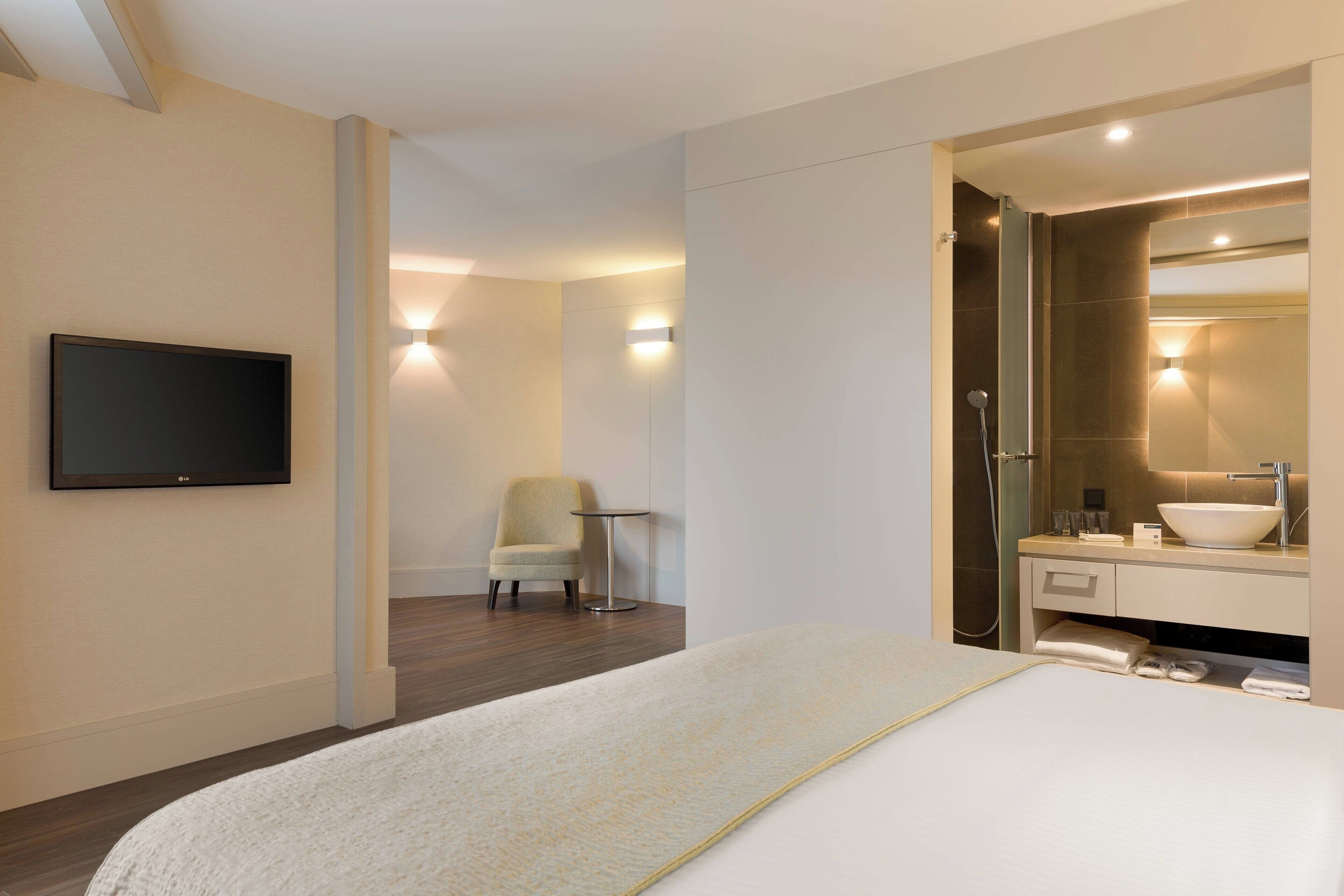 A modern hotel room with a large bed, a TV mounted on the wall, a small round table, a chair, a bathroom with a large mirror, and a sink.