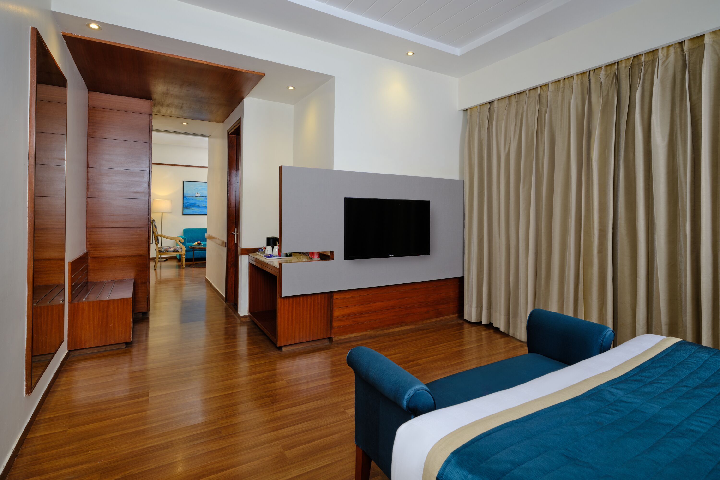 Habitación de hotel con una cama grande, televisor montado en la pared, suelo de madera y vista al pasillo.